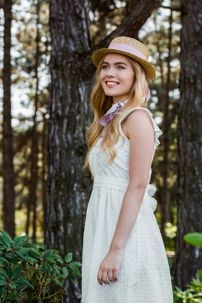 Belle jeune femme souriante en robe et chapeau en osier avec ruban debout et regardant loin dans la forêt — Photo de stock