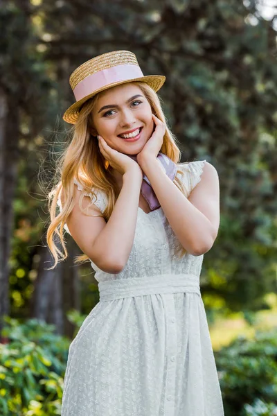 Красивая нежная блондинка в платье и плетеной шляпе с лентой улыбаясь на камеру в парке — стоковое фото