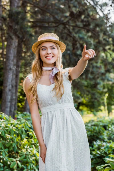 Красивая улыбающаяся девушка в белом платье и плетеной шляпе с лентой, указывающей пальцем в парке — стоковое фото