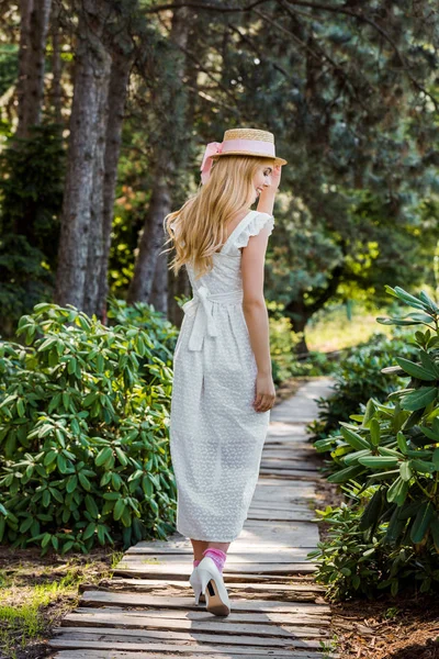Back view of beautiful girl in white dress and wicker hat walking on wooden walkway in park — Stock Photo