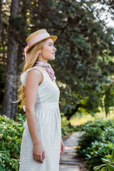 Vista lateral de hermosa mujer joven en vestido blanco y sombrero de mimbre sonriendo y mirando hacia otro lado en el parque - foto de stock