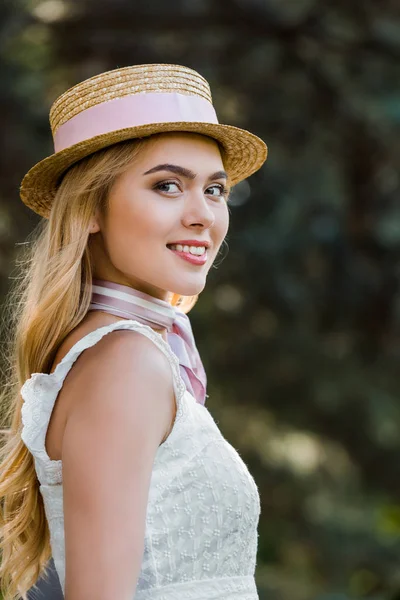 Side view of beautiful young woman in wicker hat smiling at camera in park — Stock Photo