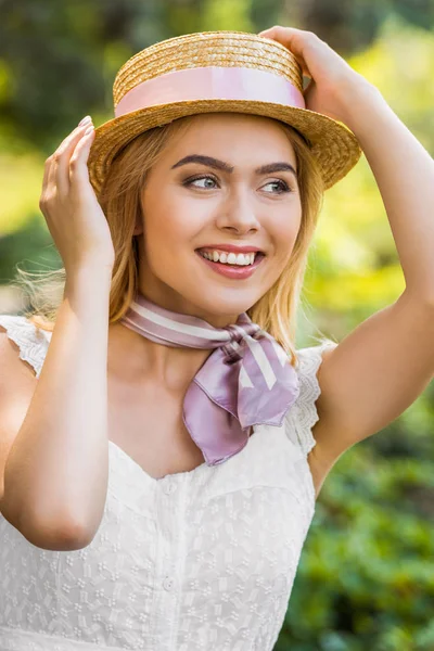 Hermosa chica rubia sonriente con sombrero de mimbre con cinta y mirando hacia fuera en el parque - foto de stock