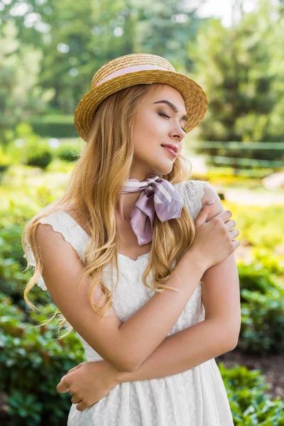 Hermosa tierna joven en vestido y sombrero de mimbre de pie con los ojos cerrados en el parque - foto de stock