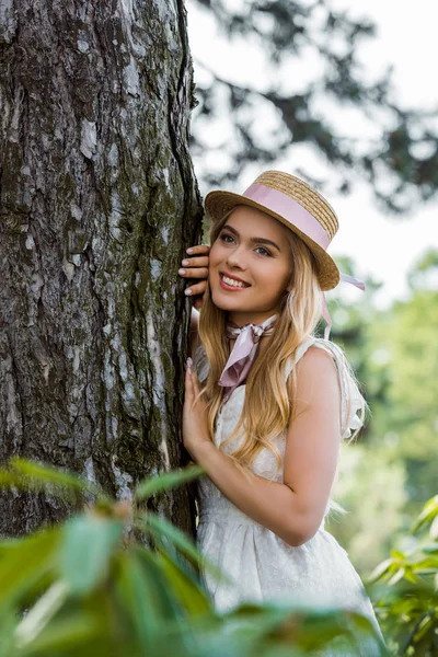 Enfoque selectivo de hermosa chica sonriente en sombrero de mimbre apoyado en el árbol en el parque - foto de stock
