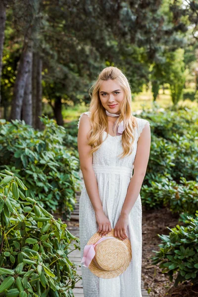 Belle jeune femme blonde en robe blanche tenant chapeau en osier et souriant à la caméra dans le parc — Photo de stock