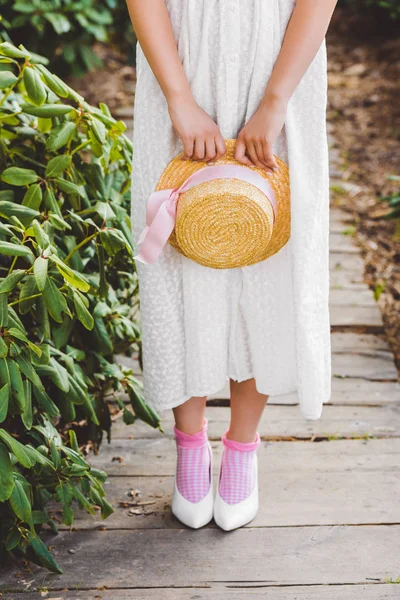 Tiro cortado de menina em vestido branco segurando chapéu de vime com fita — Fotografia de Stock