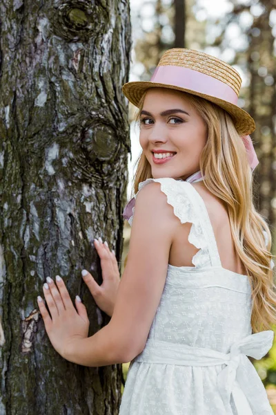 Hermosa mujer joven en sombrero de mimbre tocando el árbol y sonriendo a la cámara - foto de stock