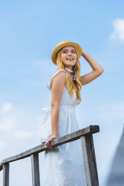 Низкий угол зрения счастливая блондинка в плетеной шляпе улыбаясь и глядя в сторону на голубое небо — стоковое фото