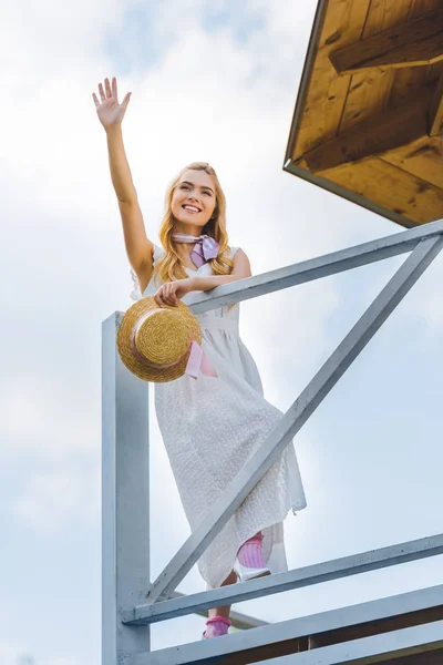 Vue à angle bas de jeune femme heureuse en robe blanche tenant chapeau en osier et levant la main — Photo de stock