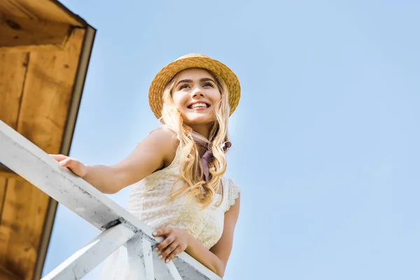 Low angle view of beautiful happy blonde girl in wicker hat leaning at railing and looking away against blue sky — Stock Photo