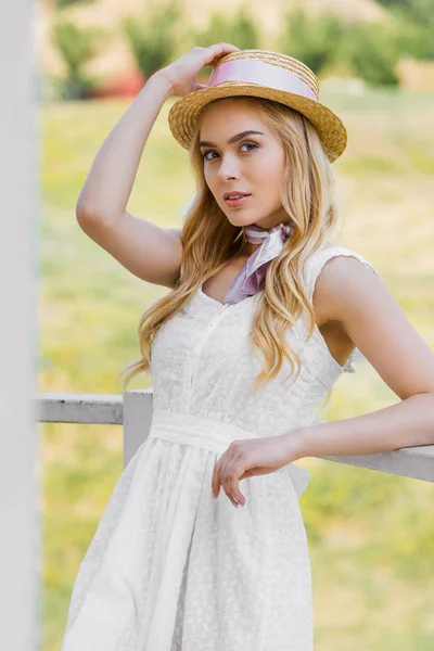 Mulher loira atraente em chapéu de vime e vestido branco inclinando-se no corrimão e olhando para a câmera — Fotografia de Stock