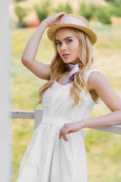 Atractiva mujer rubia en sombrero de mimbre y vestido blanco apoyado en barandilla y mirando a la cámara - foto de stock