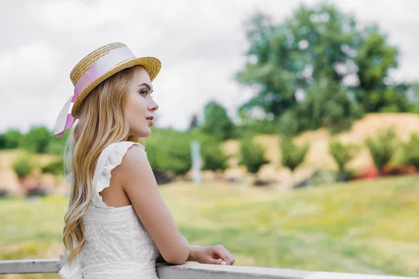 Pensive attractive blonde girl in wicker hat with ribbon leaning at railing and looking away outdoors — Stock Photo
