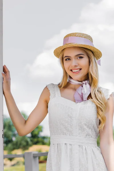 Красива молода блондинка в плетеному капелюсі і ніжне плаття посміхається на камеру — стокове фото