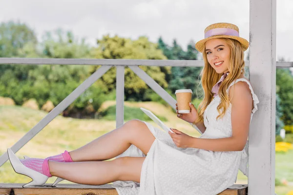 Hermosa chica en vestido blanco y sombrero de mimbre sosteniendo taza de papel y el uso de tableta digital mientras está sentado en el banco y sonriendo a la cámara en el parque - foto de stock