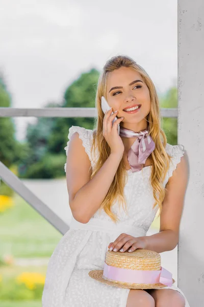 Belle femme blonde souriante tenant chapeau en osier et parlant par smartphone — Photo de stock