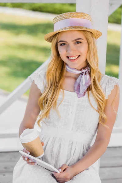 Hermosa mujer joven sonriente en sombrero sosteniendo café para ir y tableta digital mientras está sentado en el banco en el parque - foto de stock