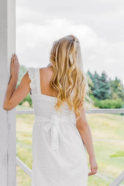 Vue arrière de belle fille blonde en robe blanche regardant les plantes vertes — Photo de stock