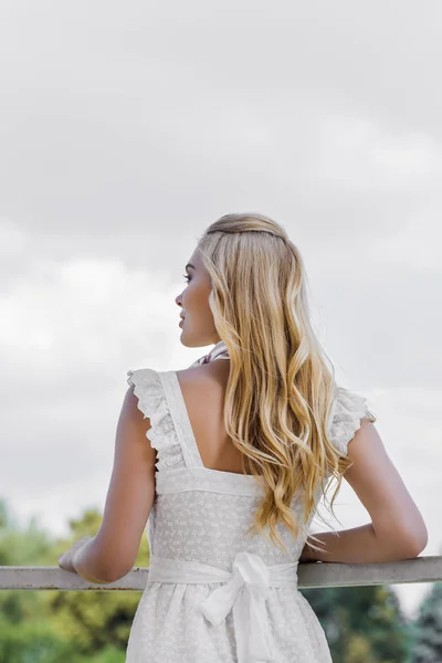 Вид сзади красивой молодой блондинки в белом платье, смотрящей вдаль в парке — стоковое фото
