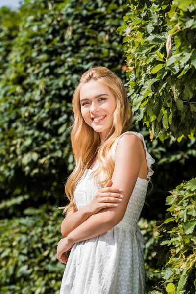 Beautiful blonde girl in white dress smiling at camera while standing near green plants — Stock Photo