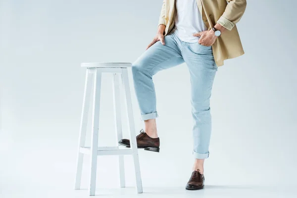 Vue recadrée d'un jeune homme élégant appuyé sur un tabouret sur fond blanc — Photo de stock