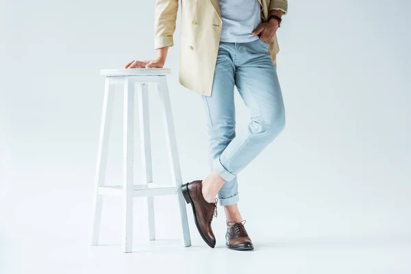 Vue recadrée d'un jeune homme élégant debout près d'un tabouret blanc sur fond blanc — Photo de stock