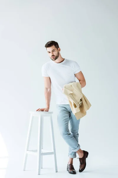 Handsome bearded man holding jacket and leaning on stool on white background — Stock Photo