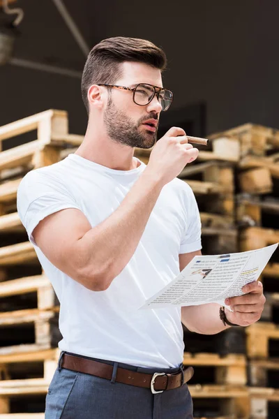 Guapo barbudo hombre de negocios en gafas de fumar cigarro mientras lee informe de negocios por paletas de madera - foto de stock