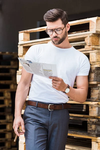 Stilvoller junger Geschäftsmann liest Geschäftszeitung und hält Zigarre an Holzpaletten — Stockfoto