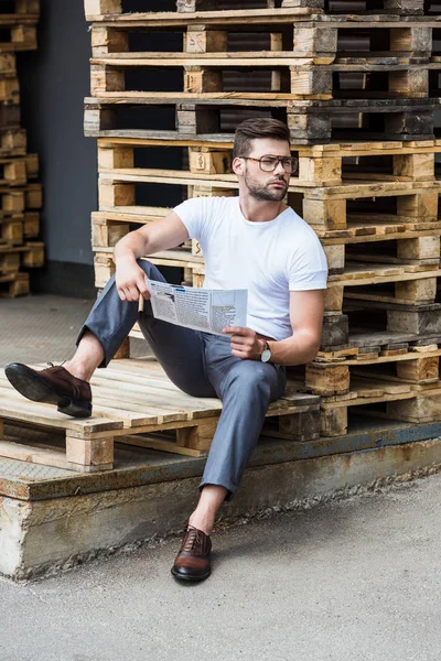 Handsome bearded businessman sitting on wooden palettes with cigar and report in hands — Stock Photo