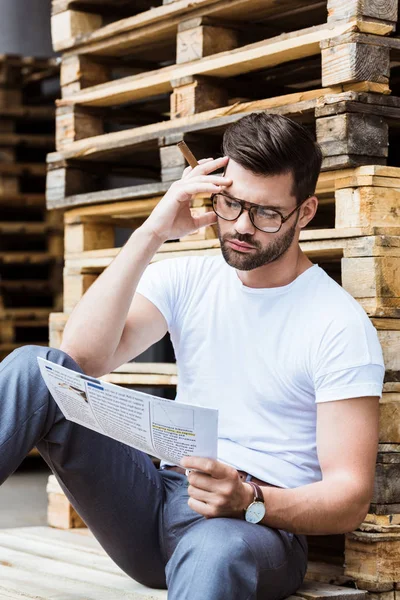 Empresario confiado de moda en gafas fumando cigarro mientras lee el documento de negocios por paletas de madera - foto de stock