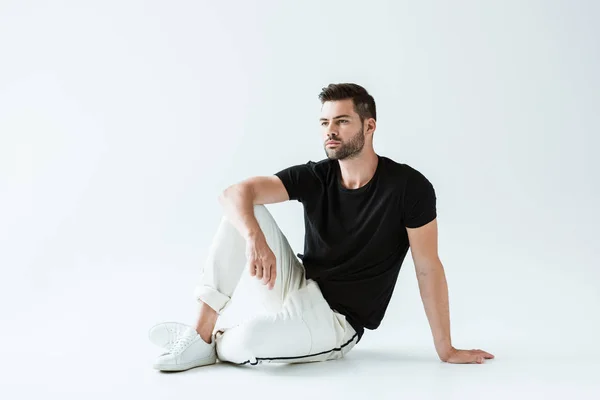 Bello uomo barbuto in abiti casual seduto sul pavimento su sfondo bianco — Foto stock