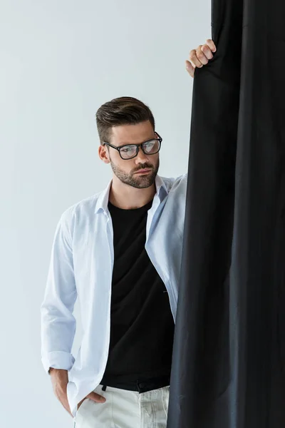 Bonito barbudo vestindo óculos e camisa branca em pé por cortina preta — Fotografia de Stock