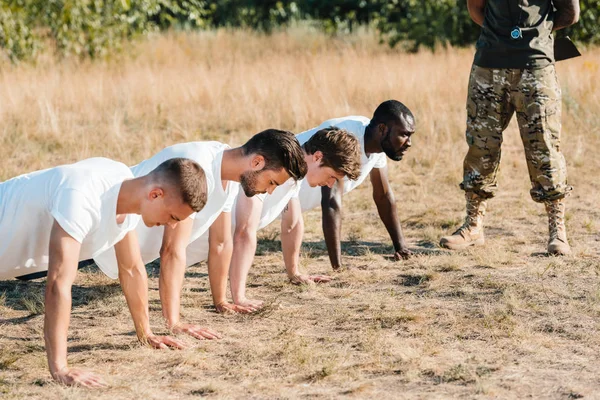 Partial view of tactical instructor examining multicultural soldiers doing push ups on range — Stock Photo