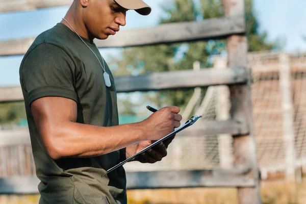 Vue latérale de l'instructeur tactique afro-américain prenant des notes dans le bloc-notes à portée — Photo de stock