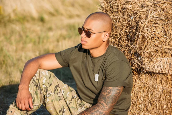 Young african american soldier in military uniform and sunglasses resting near hay on range — Stock Photo