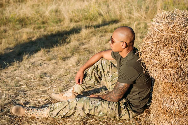 Side view of young african american soldier in military uniform and sunglasses resting near hay on range — Stock Photo