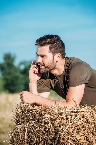 Lächelnder Soldat in Militäruniform, der auf dem Smartphone spricht, während er sich auf Heu in Reichweite lehnt — Stockfoto