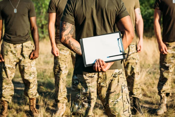 Visão parcial do instrutor tático com bloco de notas e soldados multirraciais em uniforme militar na faixa — Fotografia de Stock