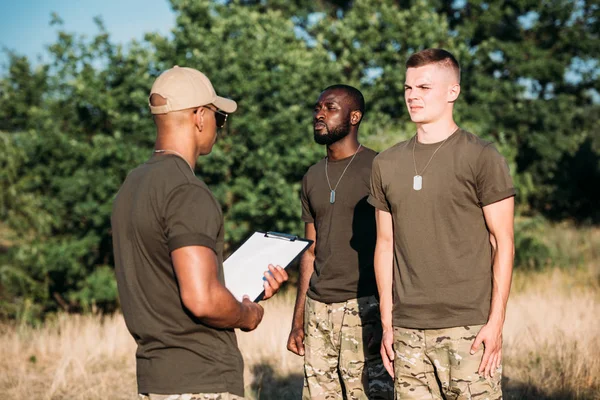 Instructor táctico afroamericano con bloc de notas y jóvenes soldados multirraciales en uniforme militar en rango - foto de stock