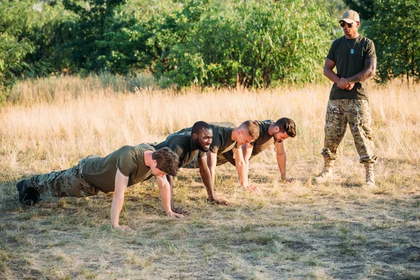 Tactical instructor in sunglasses examining multicultural soldiers doing push ups on range — Stock Photo
