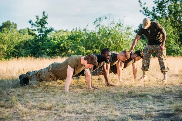 Tactical instructor examining multicultural soldiers in military uniform doing push ups on range — Stock Photo