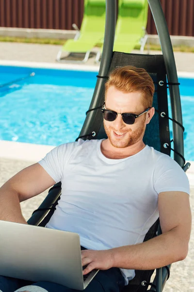 Beau jeune homme assis dans une chaise longue devant la piscine et travaillant avec un ordinateur portable — Photo de stock