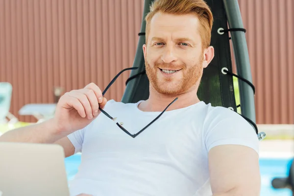 Handsome young man sitting in sun lounger in front of swimming pool and looking at camera — Stock Photo