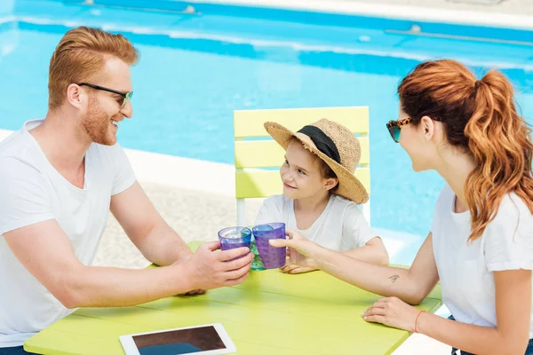 Hermosa familia joven tintineo gafas mientras está sentado en frente de la piscina - foto de stock
