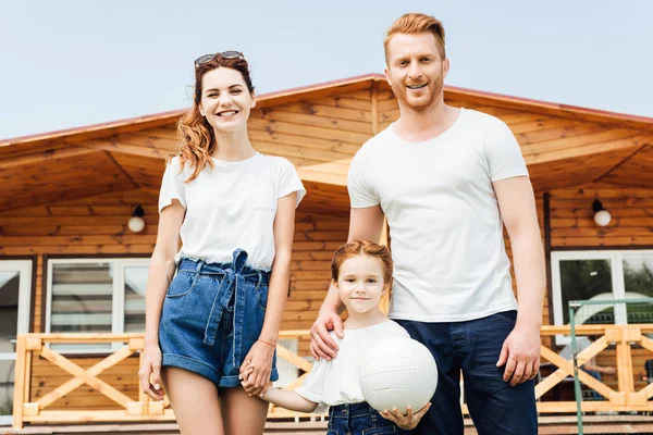 Bela família jovem com bola de vôlei em pé na frente da casa de campo de madeira — Fotografia de Stock
