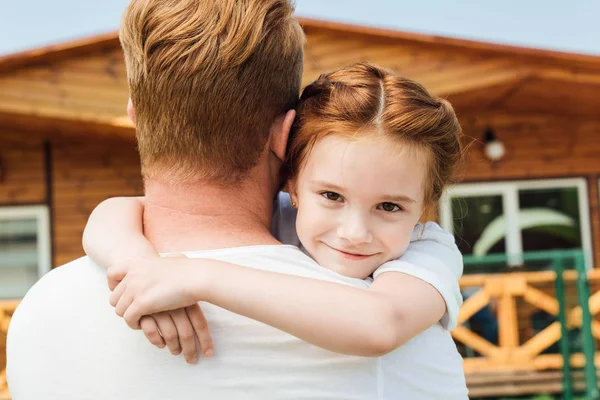 Rückansicht des Vaters, der seine entzückende kleine Tochter umarmt, während sie in die Kamera blickt — Stockfoto