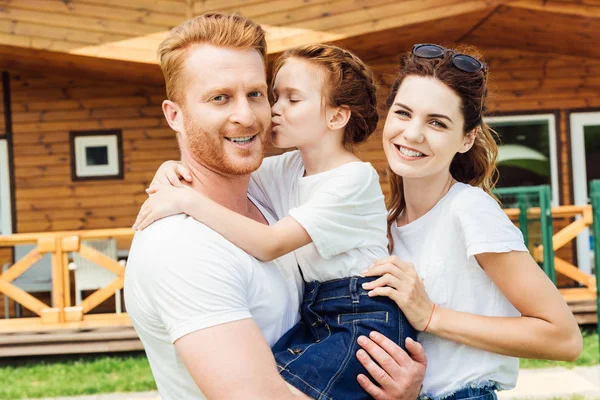 Familia joven y feliz abrazo en el jardín de casa de madera y mirando a la cámara - foto de stock