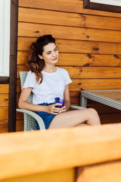 Beautiful young woman with glass of drink sitting on chair on terrace of wooden cottage — Stock Photo